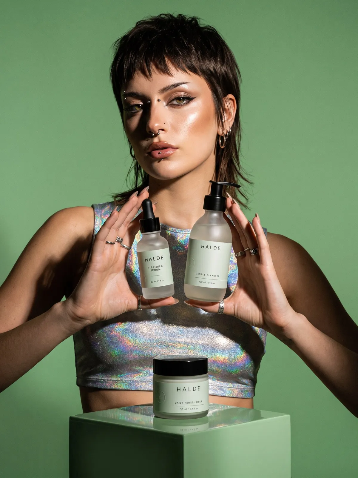 A model in a silver crop top holds HALDE bottles above a sage plinth