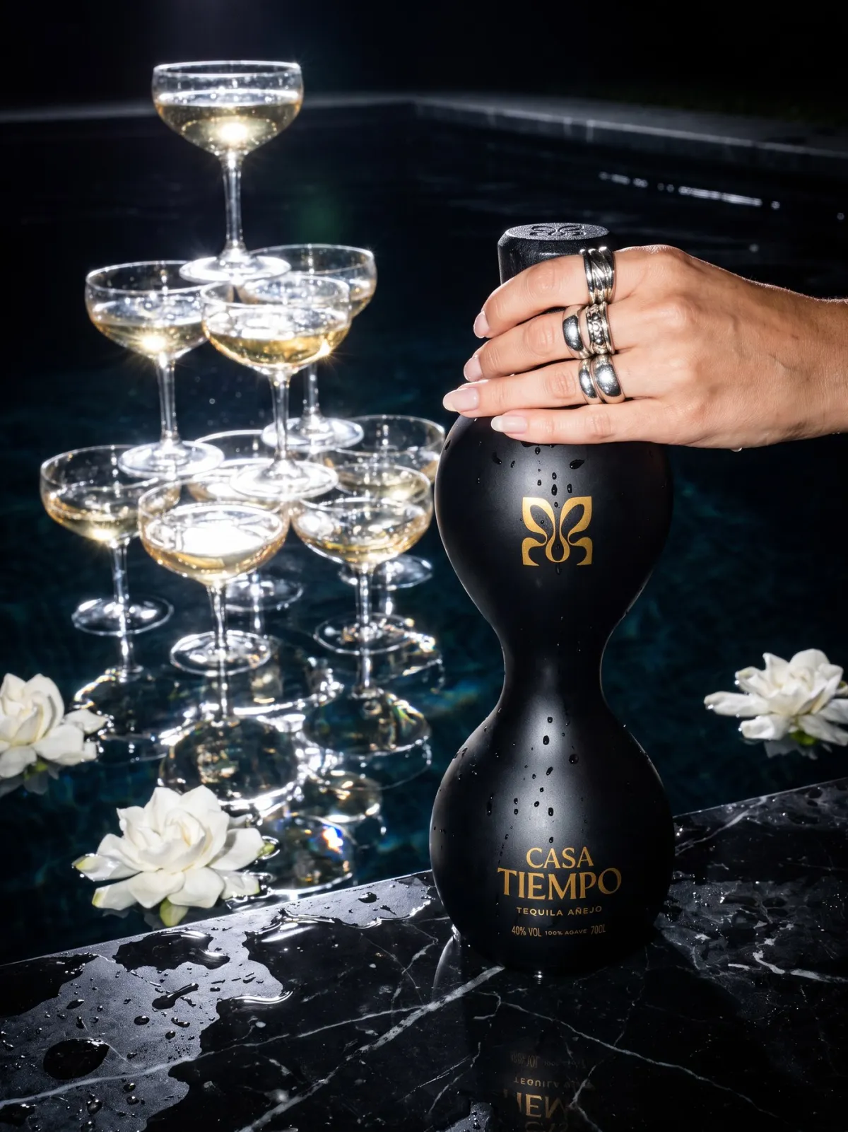 A ringed hand on a black Casa Tiempo añejo bottle beside a coupe tower