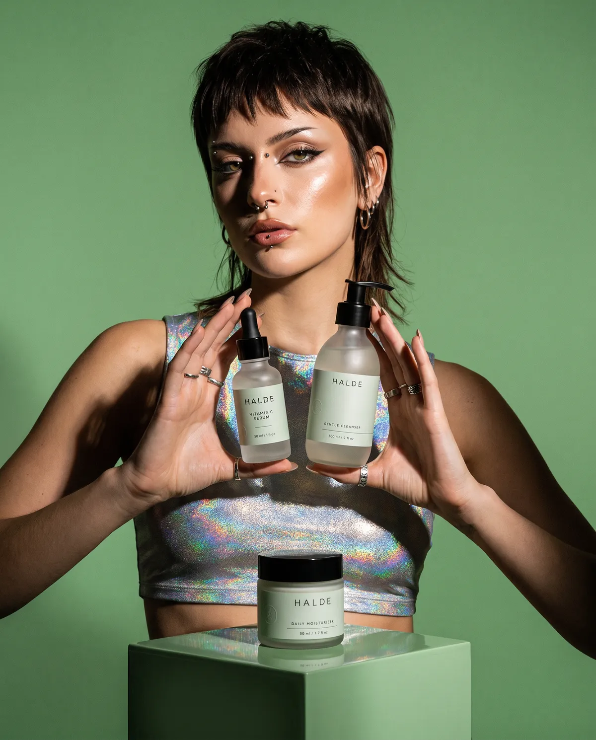 A model holds the HALDE serum and cleanser at chest height, with the moisturiser on a sage plinth in front of her.